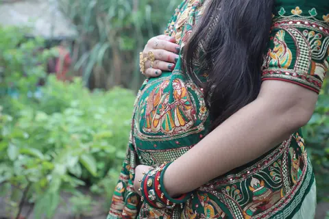 Close-up of a pregnant woman's belly in an embroidered green traditional outfit, her hand resting on top, set against a garden background.